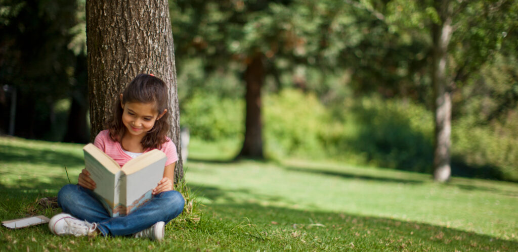 A young girl sitting on the grass beneath a tree happily reading a book. 