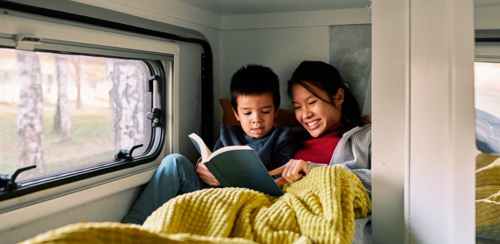 A mom and a son sitting with a cozy blanket in their camper reading a book. 